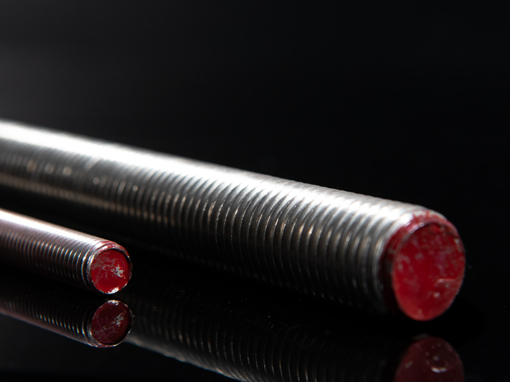 Zwei Edelstahl-Gewindestangen in unterschiedlichen Durchmessern mit roten Endkappen, liegend auf schwarzer Fläche, scharfe Detailaufnahme der Gewinde