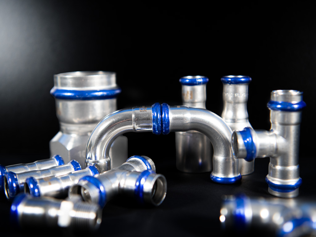 erschiedene Edelstahl-Pressfittings mit blauen Dichtungen, darunter Rohrverbinder, Bögen und T-Stücke, glänzend auf schwarzer Fläche präsentiert