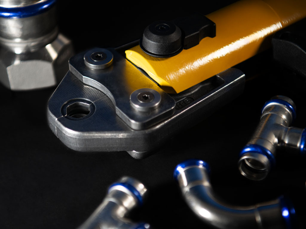 Elektrisches Presswerkzeug mit gelbem Griff zum Verpressen von Edelstahl-Pressfittings, umgeben von T-Stücken und Rohrverbindern