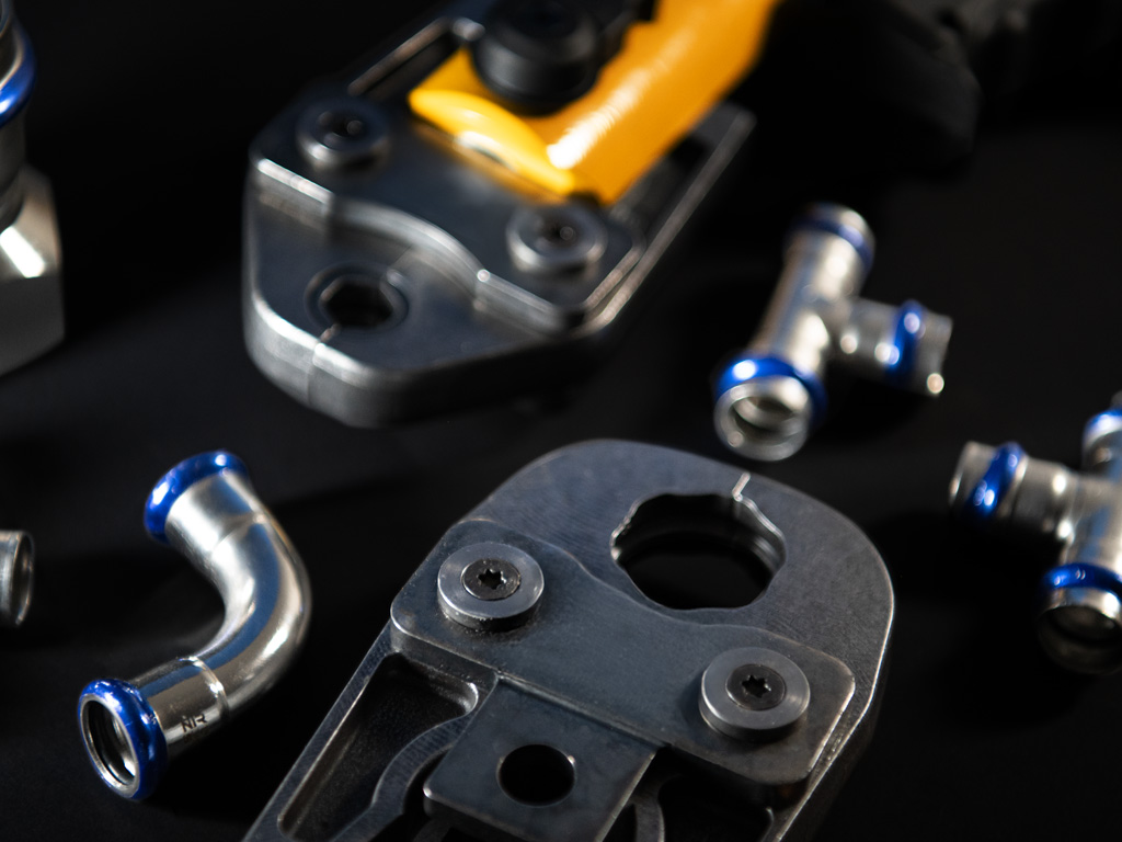 Nahaufnahme einer Pressbacke aus Metall für Presswerkzeuge, umgeben von Edelstahl-Pressfittings und Rohrverbindern auf schwarzem Hintergrund
