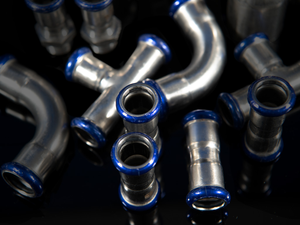 Edelstahl-Pressfittings mit blauen Dichtungsringen in verschiedenen Formen, darunter Bögen und T-Stücke, auf schwarzem Hintergrund