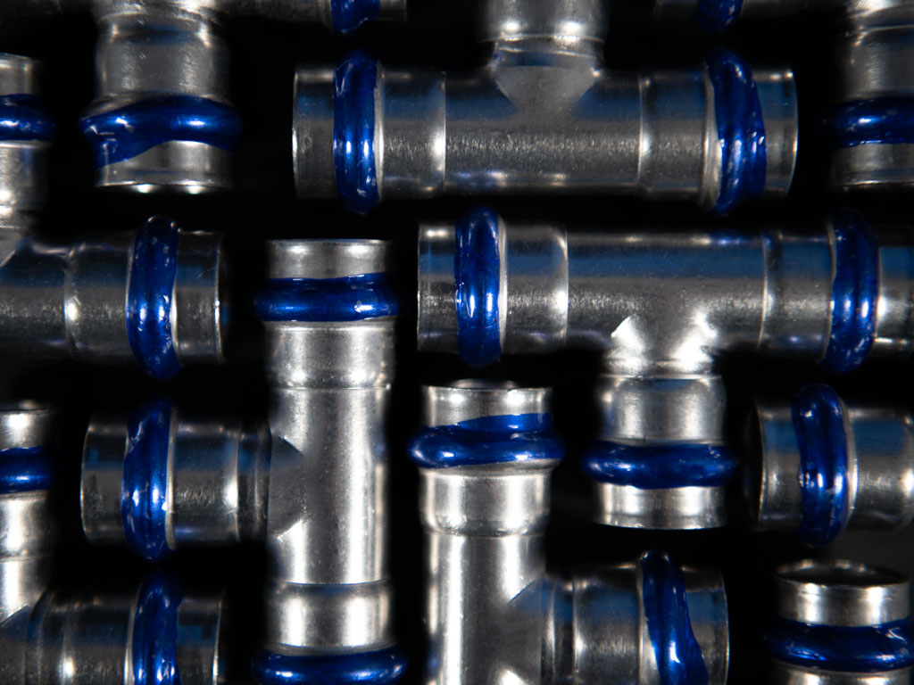 Mehrere Edelstahl-T-Stücke und Rohrfittings mit blauen Dichtungsringen in dichter Anordnung, gleichmäßig beleuchtet